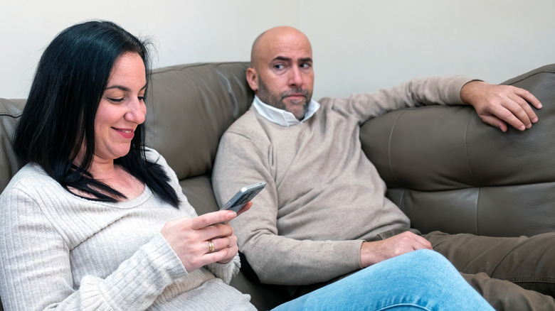 A woman smiles while using her smartphone on a sofa, as a man beside her looks on with a serious, uneasy expression.