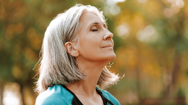 A serene older woman is standing outdoors with her eyes closed