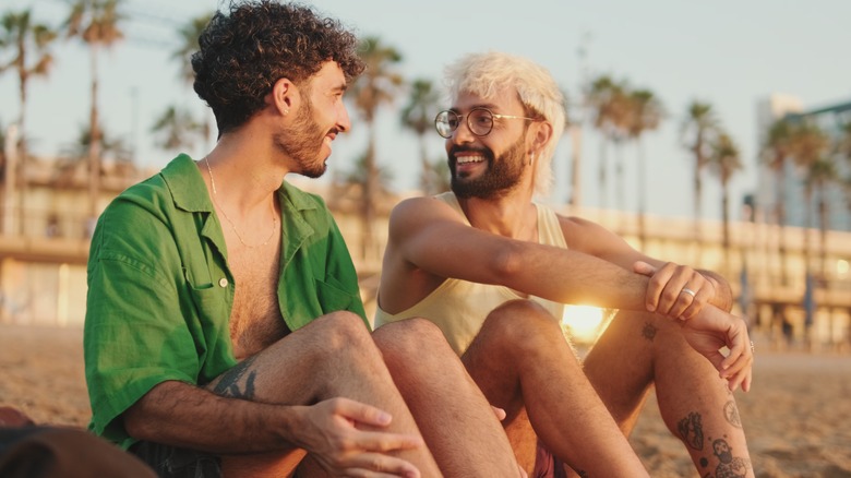 gay couple in love talking and laughing while sitting on the beach at dawn