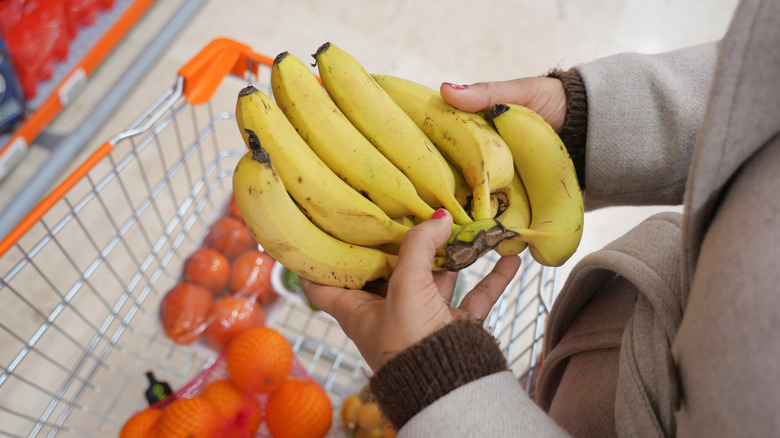Person holding bananas over a shopping cart full of other fruit