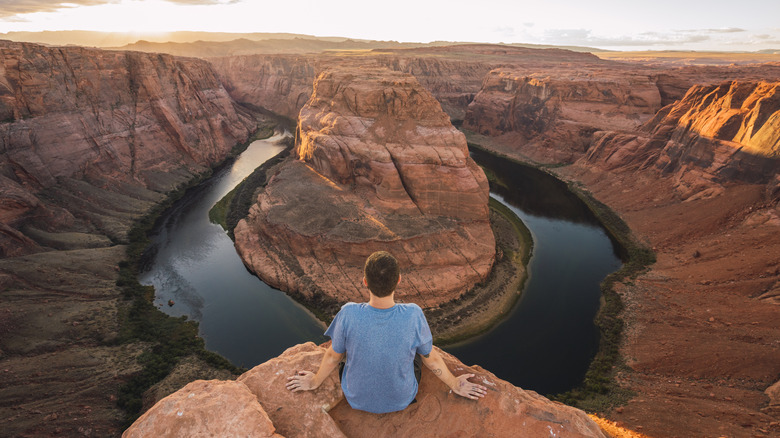 Man in blue shirt sits on ridge of Grand Canyon, river below