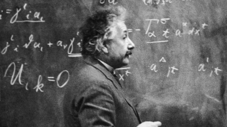 Albert Einstein with greying hair and mustache stands in front of a blackboard covered in equations