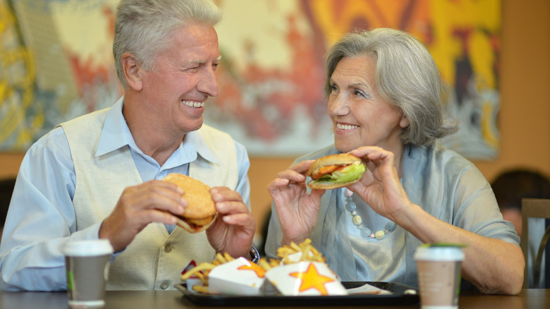 Grey-haired older couple smiling and sitting side by side with burgers