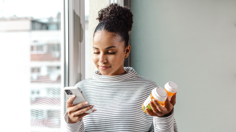 Young woman smiles while looking at phone in one hand and holding pill bottles in other hand