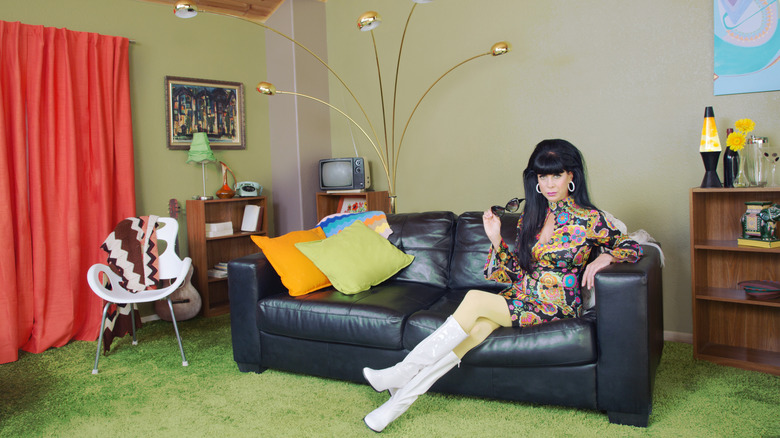 Woman in colorful dress and white boots sits on black sofa in a vintage style living room