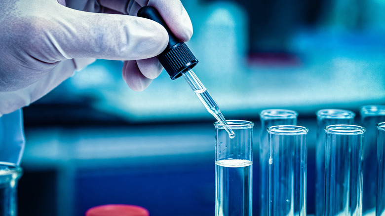 A gloved hand uses a dropper to dispense a liquid into a tube.