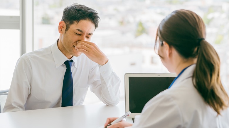 Man in tie plugs his nose with his hand, sitting across from a nurse with a laptop