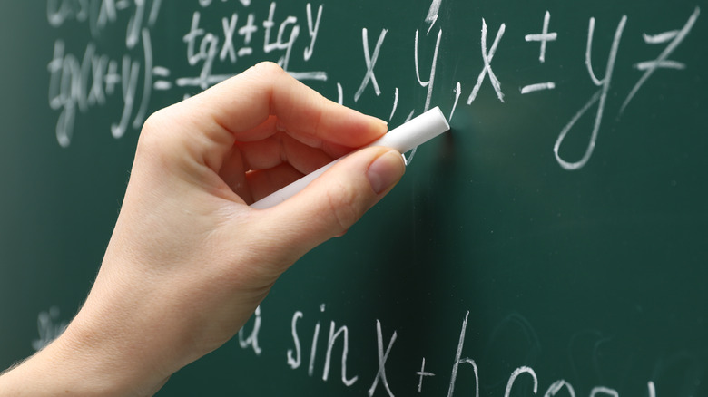 A close-up of a hand writing math equations on a chalkboard.