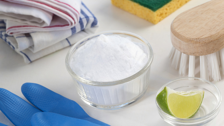 Small bowl of baking soda surrounded by gloves, cleaning tools, and a lime wedge