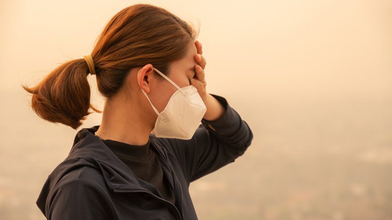 Woman wearing a mask and holding her head as wildfire smoke hides the city in the background