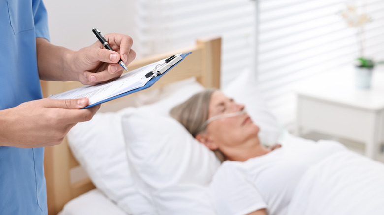 A doctor's hands hold a clipboard with an unconscious mature woman in a hospital bed in the background