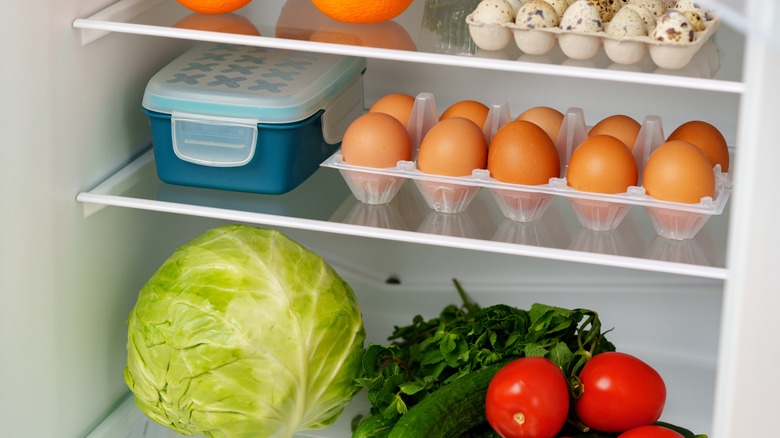 A white refrigerator is open, revealing a well-stocked interior with fresh fruits, vegetables, eggs, and other groceries