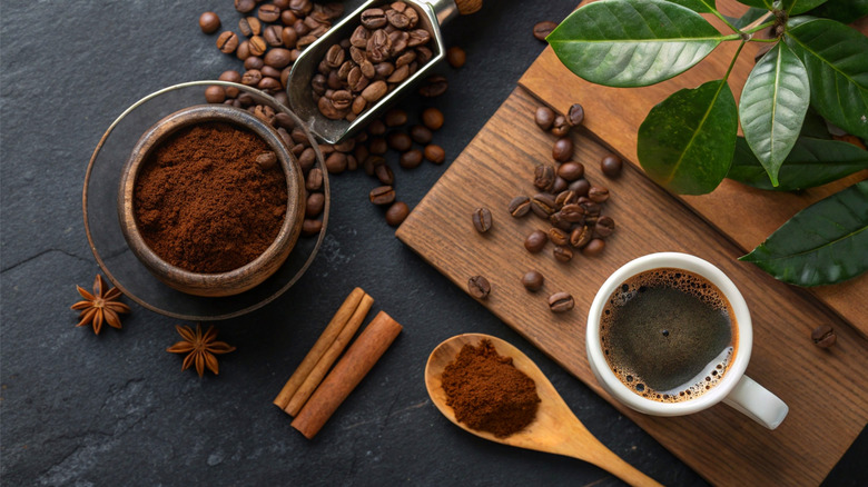 Freshly ground coffee, whole coffee beans, and a steaming cup of coffee sit on a textured slate background