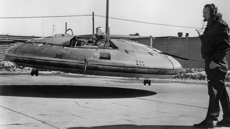 Pilot in Avrocar flying-saucer-shaped craft rises above the ground at a slight angle