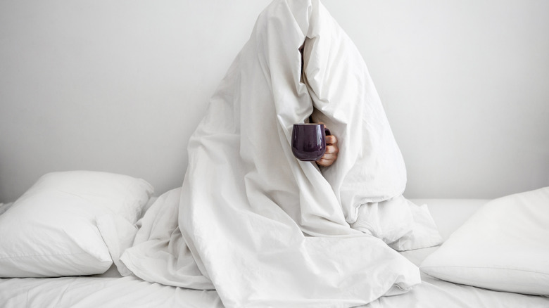 Person holding coffee mug in bed with blanket over head