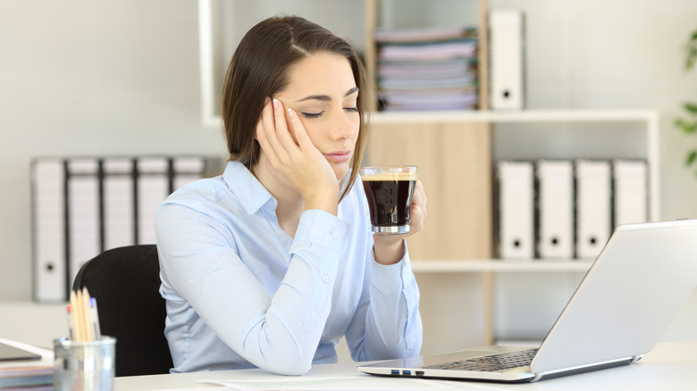 Woman with eyes closed in front of laptop while holding cup of coffee