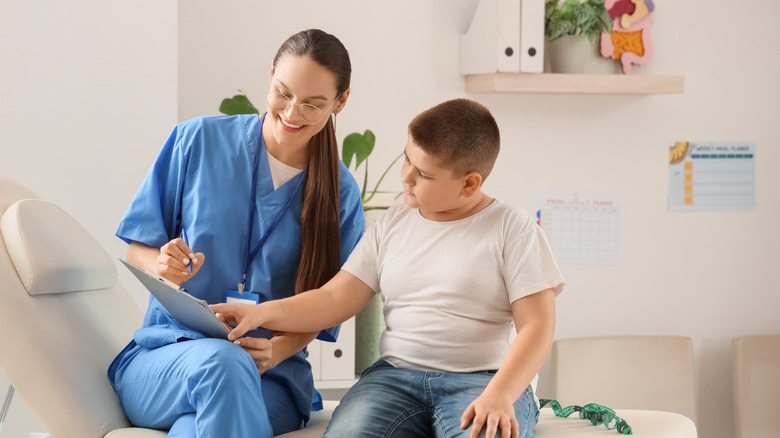 Young chubby boy sits on examining table while female nurse points to a chart