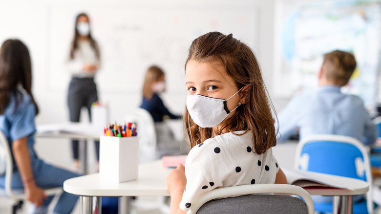 Young girl seated in class with mouth covering, teacher, also masked, in background