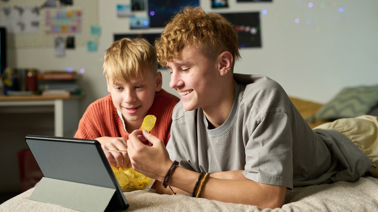 Smiling teenager and his young brother eating chips and watching laptop