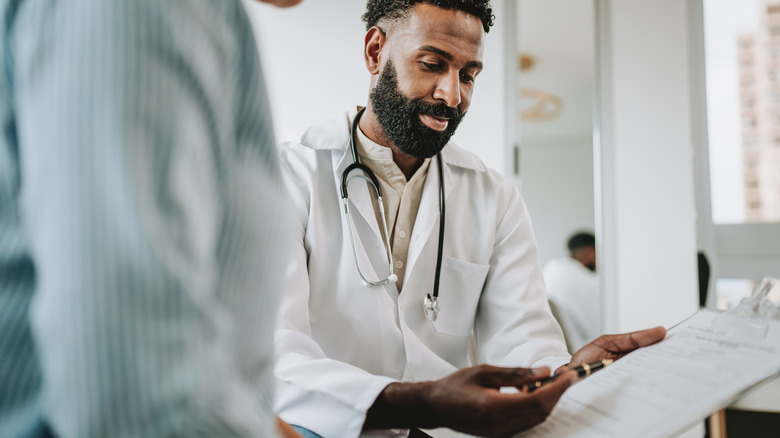 A doctor holds a clipboard and points to it with a pen while speaking to a patient who is blurred in the foreground.