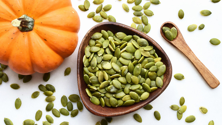 pumpkin seeds in a wooden bowl and wooden spoon next to a small orange pumpkin