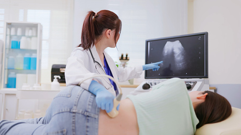 A woman lies on her side as a nurse passes an ultrasound wand over her and points to a screen