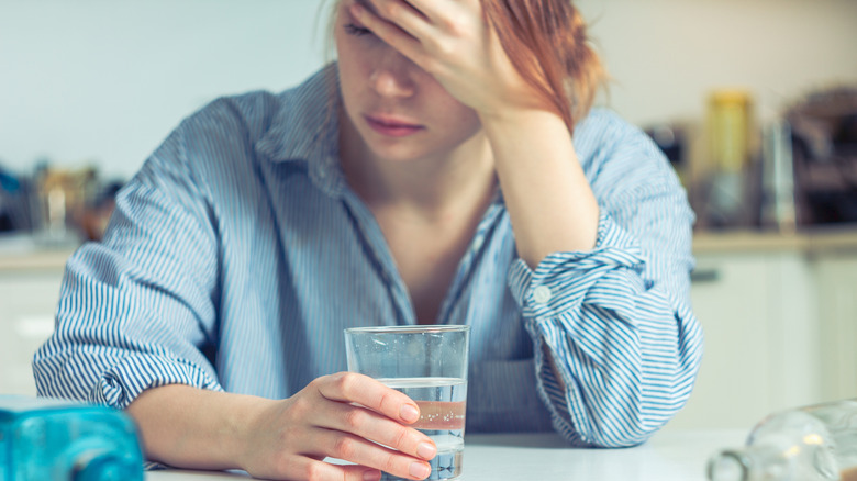 Woman sitting at a table with a glass of water, holding her head as if having a headache