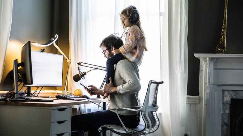 A man works at a desk while a small girl sits on his shoulders