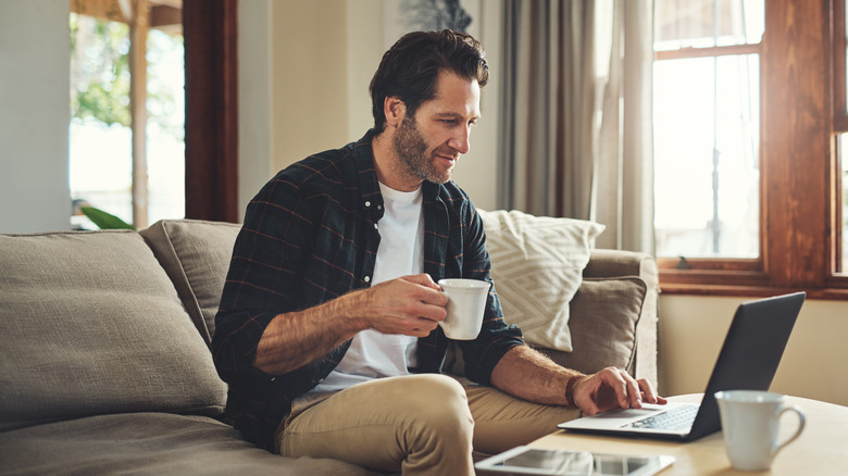 A man sits in his living room holding a mug and working on a laptop