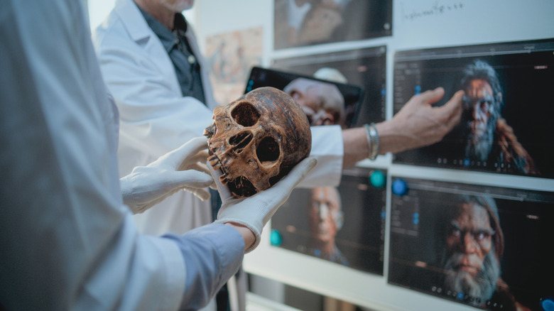 Gloved lab worker holds human skull with screens in background