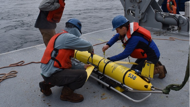 Two technicians with lifejackets apply tools to a torpedo-shaped instrument