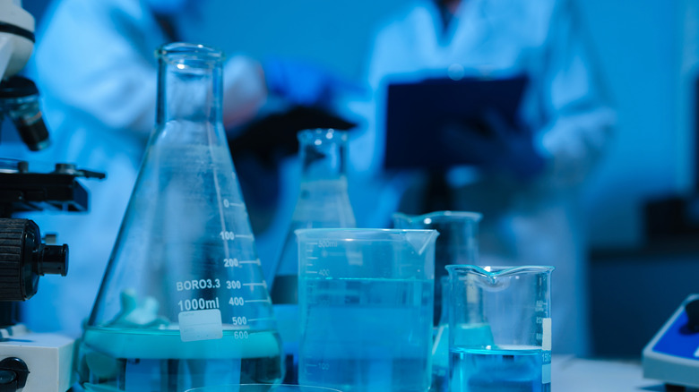 A close-up of several flasks with scientists conversing in the blurred background.
