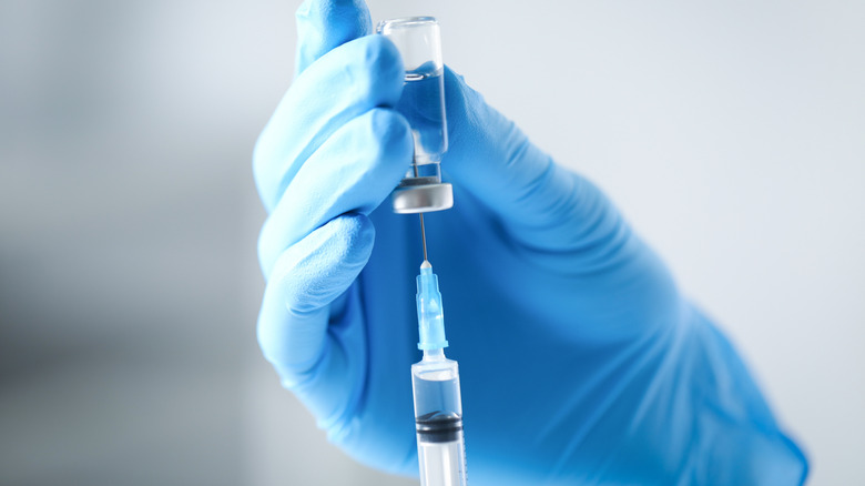 Gloved hands using a syringe to withdraw a solution from a glass vial.
