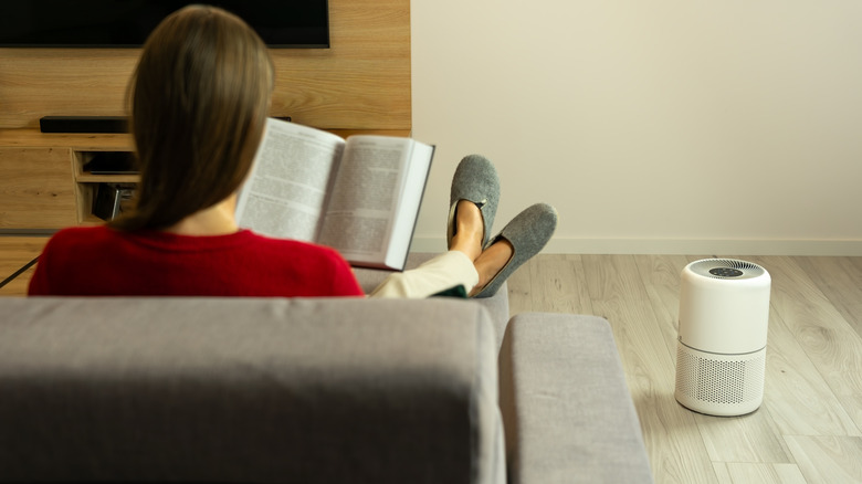 A woman in a red shirt sits on a sofa reading a book while an air purifier sits on the floor next to her