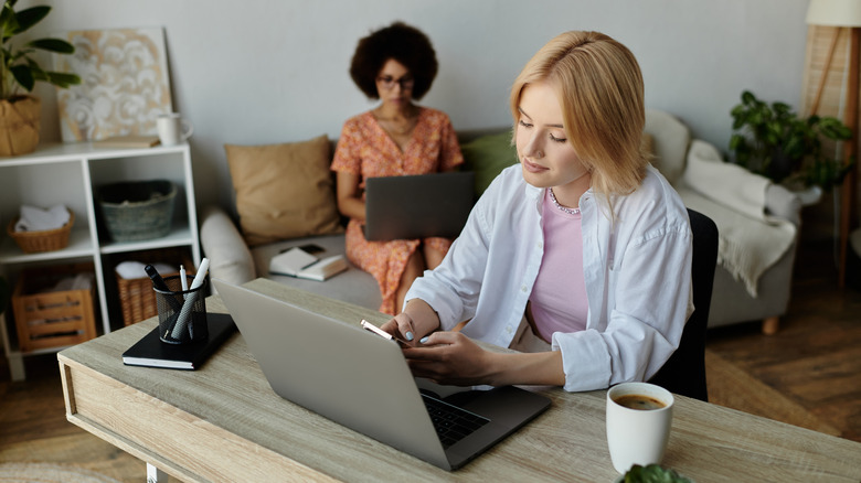 A lesbian couple works from home, one on a laptop and the other on her phone