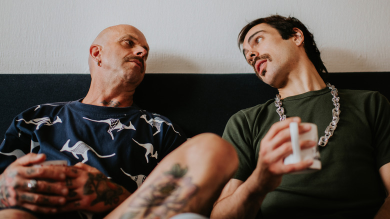 A gay couple look at each other as they sit against a headboard on a bed