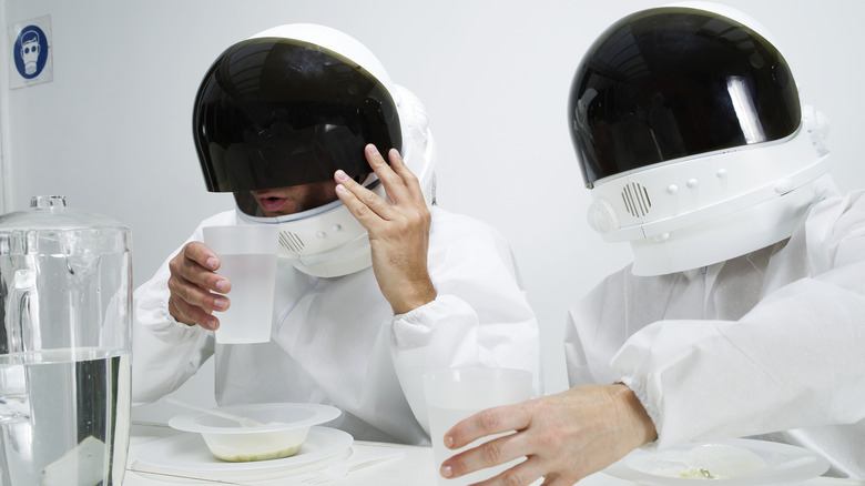 Two people wearing white space suit costumes with helmets sitting at a table. One is lifting helmet visor to drink a glass of water.