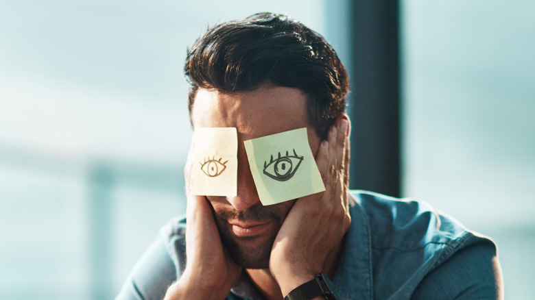 Businessman falling asleep at his desk with sticky notes over his eyes with open eyes drawn them