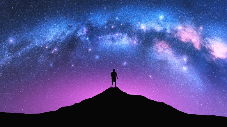 Milky Way arch and man on the mountain peak at starry night. Silhouette of alone guy, pink sky with bright stars in summer.