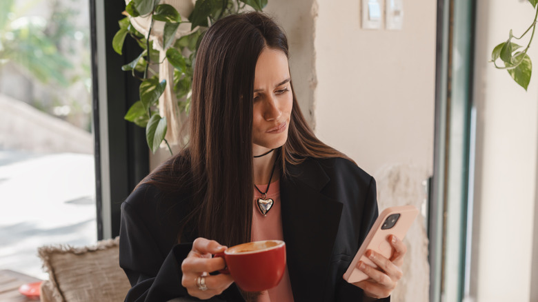 Woman with a cup in one hand in phone in the other looking puzzled