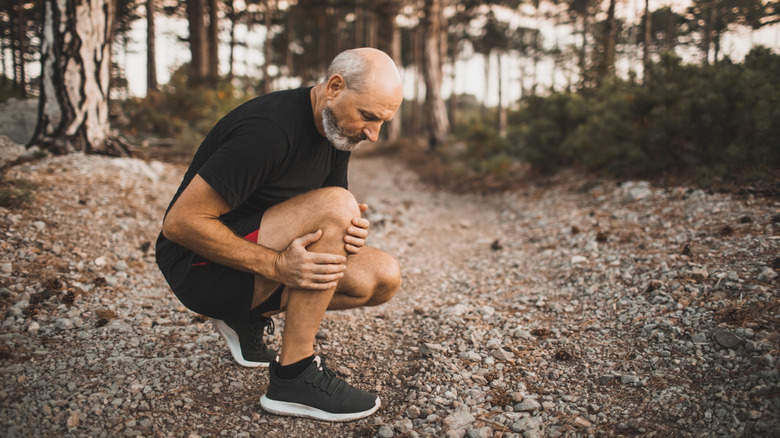 older bald man crouched rubbing knee injury on forest trail