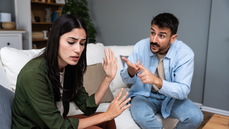 Man and woman arguing, woman raising hands in protest, man pointing to his hands with frustrated expression