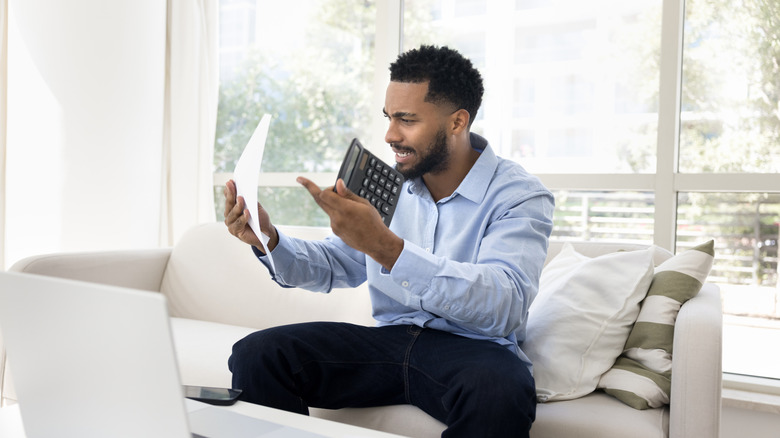 Angry professional man holding calculator and piece of paper while sitting on white couch