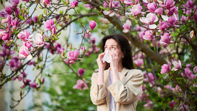 A woman with dark hair in a tan jacket blowing her nose while standing under a tree with pinkish blossoms