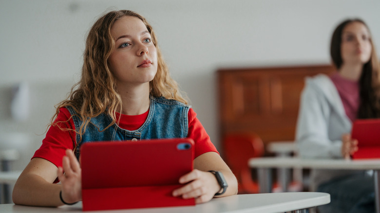 A student sits at a desk, holding an iPad and looking beyond the camera.
