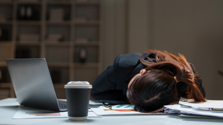 Someone sleeping on a desk beside a laptop and a cup of coffee.