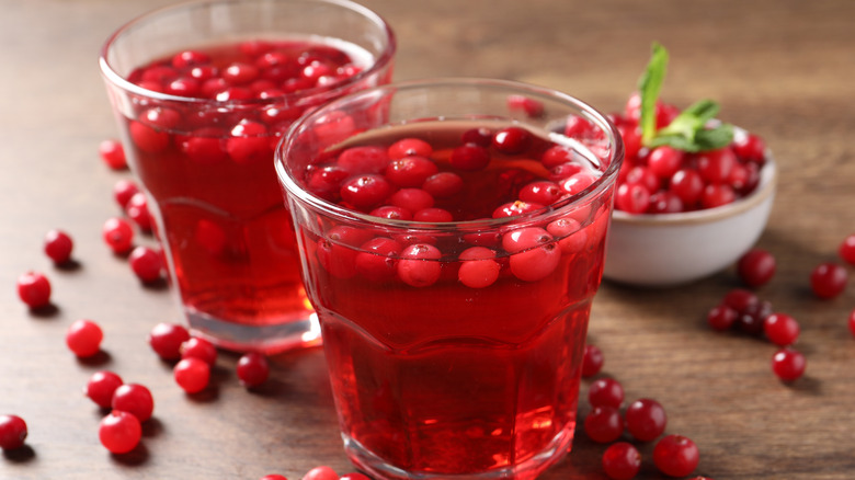 Two cups of cranberry juice on a table with cranberries surrounding them and floating in the cups while a bowl of cranberries rests blurred in the background.