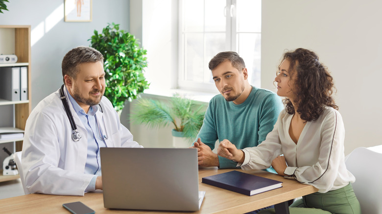 Doctor in white coat sits with a male patient and female patient discussing information on a laptop screen