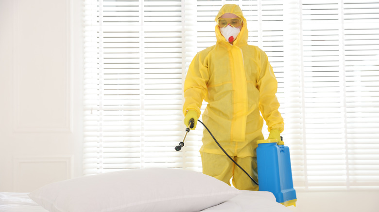 Pest control worker wearing a mask and yellow protective suit spraying pesticide near a bed
