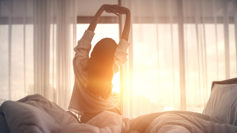 A woman stretches on her bed in front of a window as the sun rises.